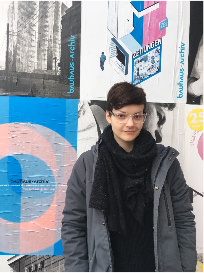 Samantha wears a black and grey outfit and is standing alongside a wall with colorful posters. Caption: At the Bauhaus Archiv in Berlin during my time living in Rostock. Photo by Mary Lawrence.